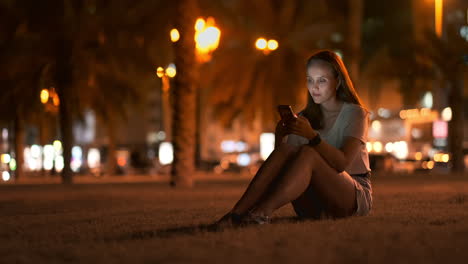 Nachts-Sitzt-Ein-Junges-Mädchen-Im-Gras-Einer-Großstadt,-Schaut-Auf-Den-Smartphone-Bildschirm,-Schreibt-Eine-Nachricht-Und-Pflegt-Ihr-Persönliches-Blog.